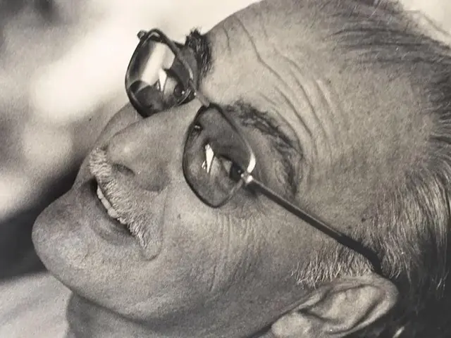 The image shows a black and white photo of a man with glasses, smiling, with a blurred background....