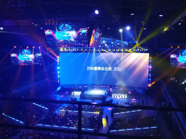 The image shows a large crowd of people sitting in front of a stage, illuminated by bright lights...