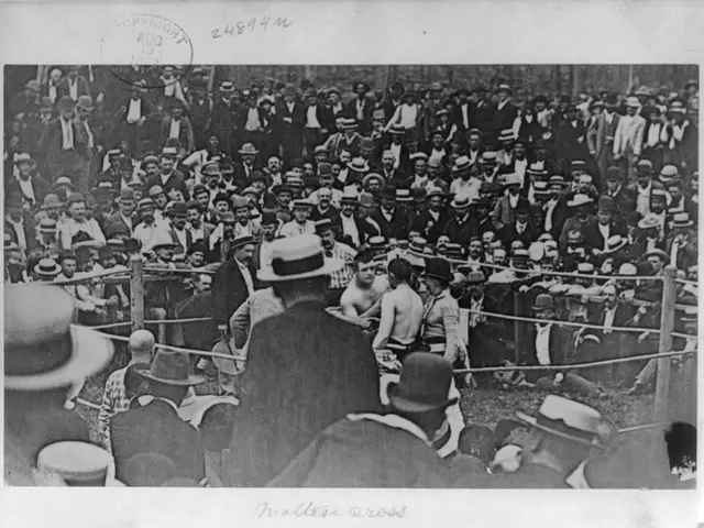 The image shows a black and white photo of a crowd of people in a boxing ring, with some of them...