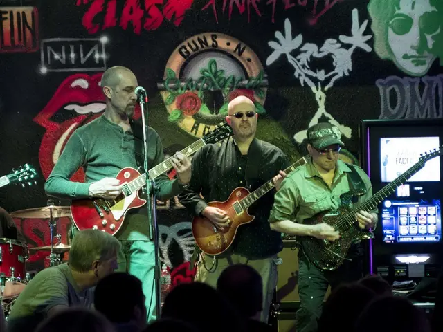 This image is clicked in a concert. There are three men in this image. At the bottom, there is a...