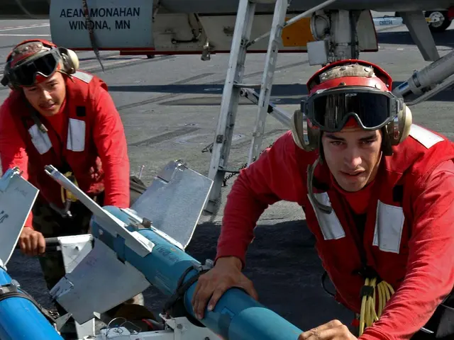 The image shows two men in red uniforms working on a missile on an aircraft carrier. They are...