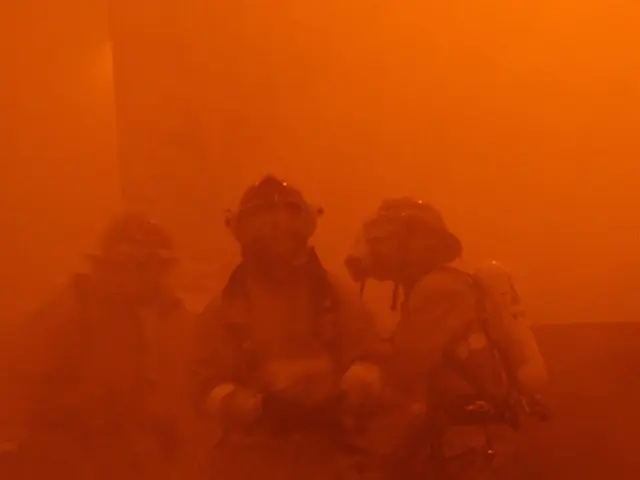 The image shows a firefighter laying on the ground in front of a raging fire, surrounded by a group...