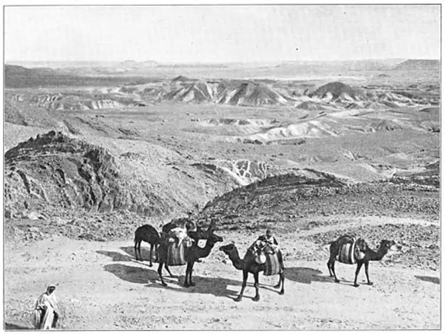 The image shows a group of people riding on the backs of camels in the desert, with hills in the...