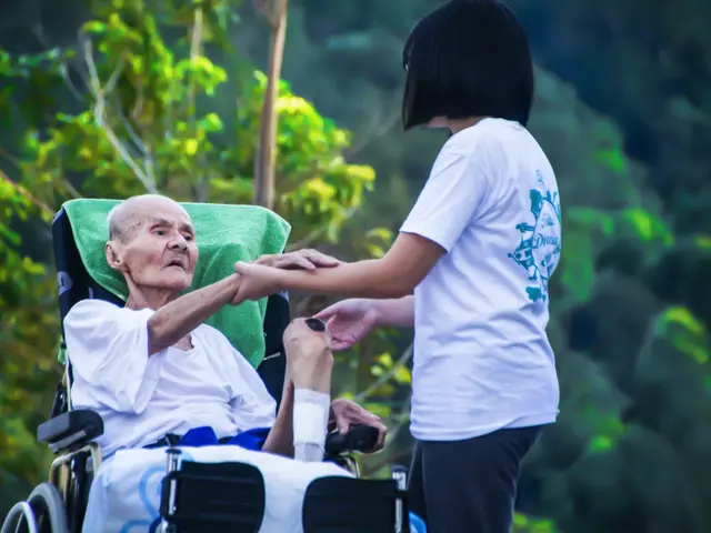 The image shows an elderly man in a wheelchair being assisted by a woman wearing a white t-shirt...