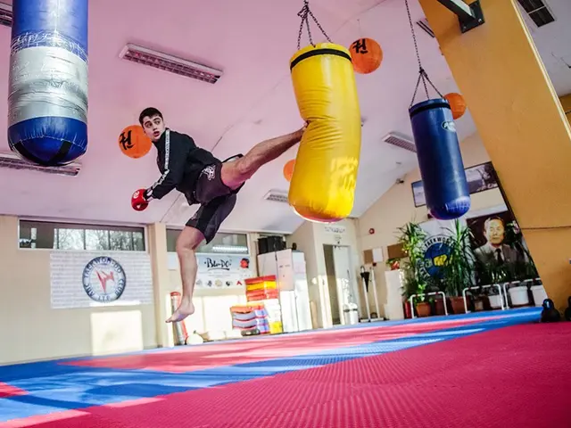 The image shows a man in mid-air, kicking a punching bag in a gym. The gym is filled with punching...