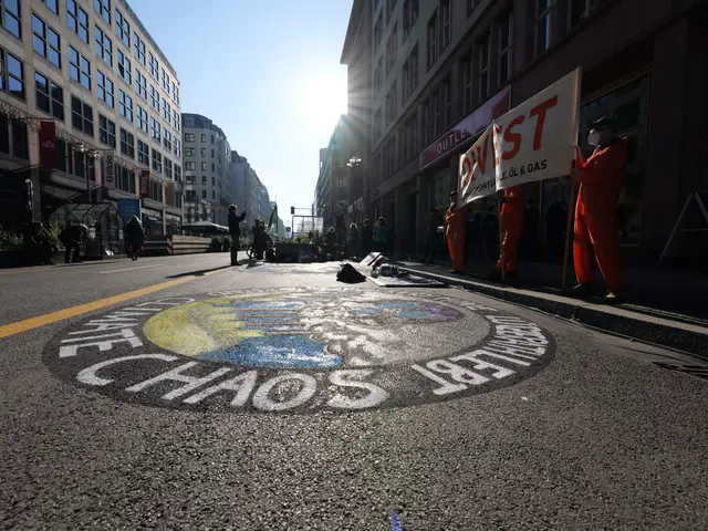 The image shows a group of people standing on the side of a street, holding banners and placards...