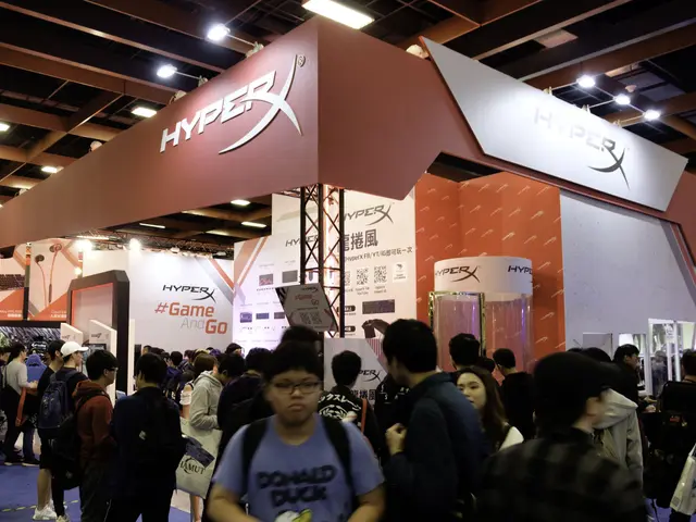 The image shows a large group of people standing in front of a booth at a gaming expo. Many of the...