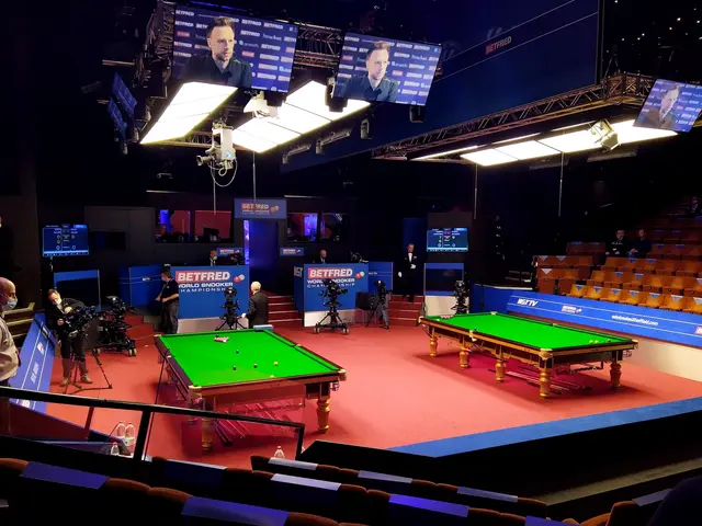 The image shows a large room with a snooker table in the middle of it, surrounded by people playing...
