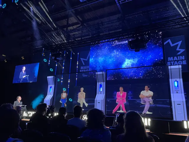 The image shows a group of people sitting on top of a stage, illuminated by bright lights and...
