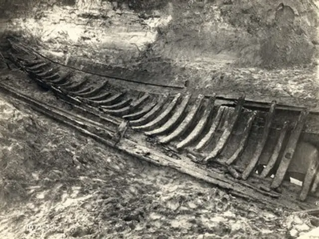 The image shows an old black and white photo of a wooden boat in the mud, with a wall in the...