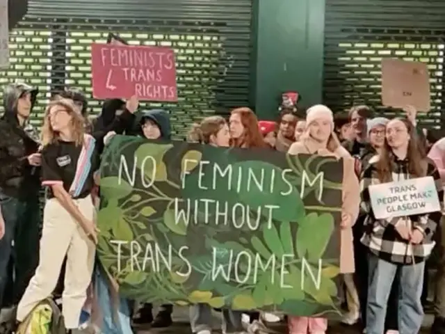 The image shows a group of people standing on the ground, holding a banner that reads "No Feminism...
