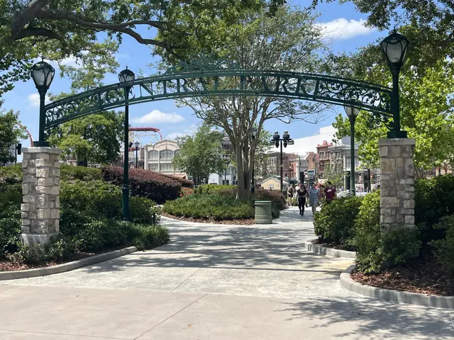 The image shows an archway in the middle of a park with people walking through it, surrounded by...