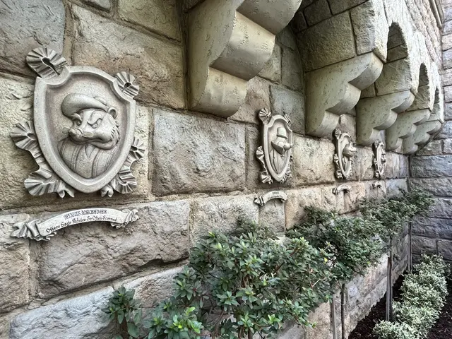 The image shows a stone building with a stone wall featuring a lion head on it, surrounded by...