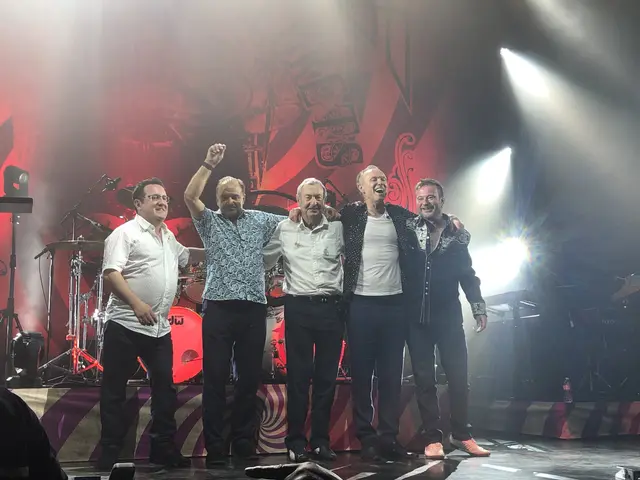 The image shows a group of men standing on top of a stage, smiling and surrounded by musical...