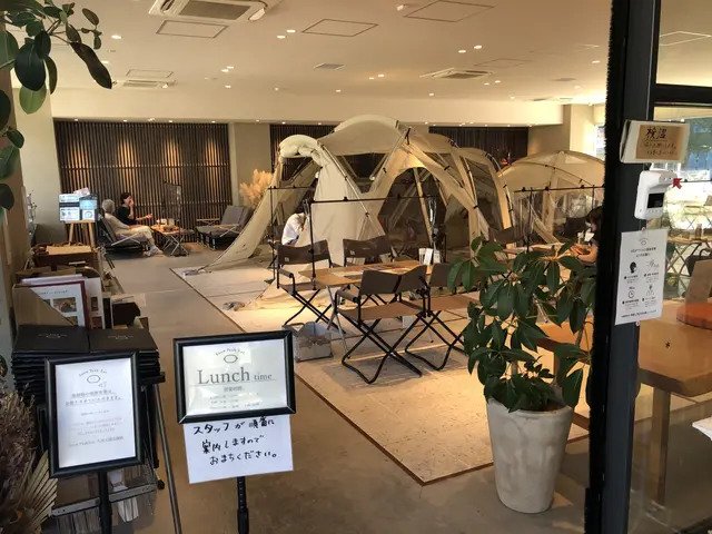 The image shows a restaurant with tables and chairs in front of a glass door, surrounded by plants,...