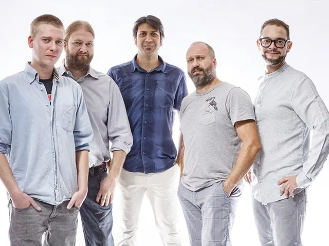 The image shows a group of men standing next to each other in front of a white background, with one...