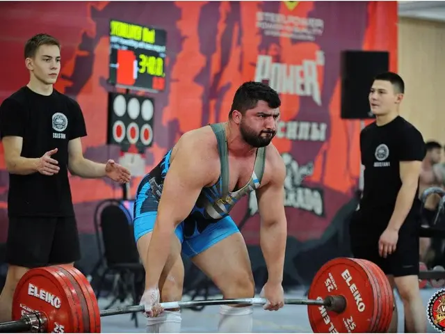 The image shows a man lifting a barbell in a weightlifting competition, surrounded by a group of...
